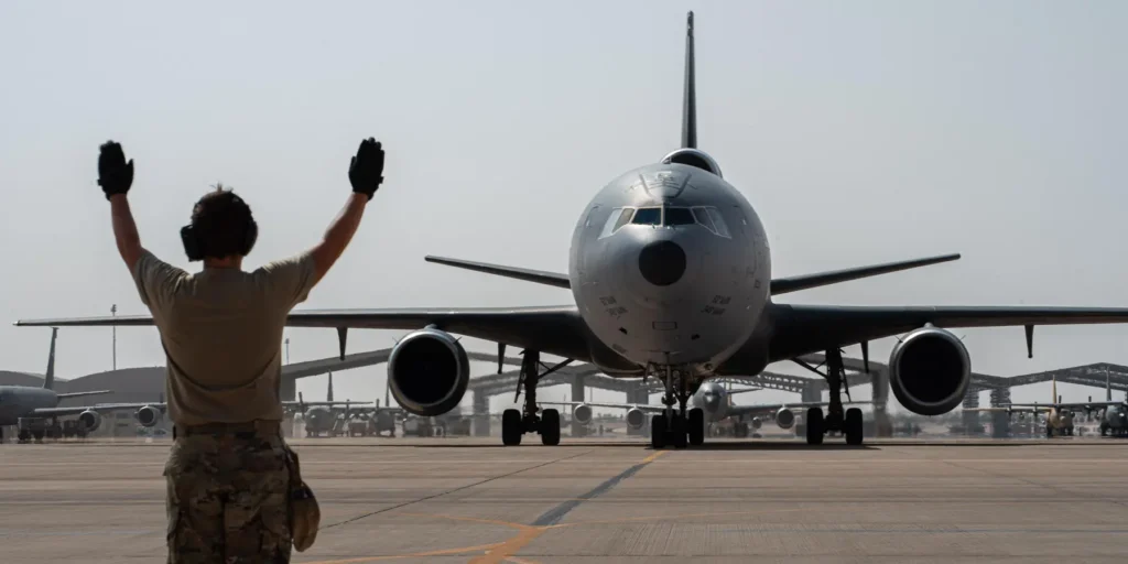 A photograph showing a US Air Force ground crew member in camouflage fatigues and hearing protection, raising both black-gloved hands to marshal a large grey military tanker aircraft taxiing on an airfield, with other military planes visible in the hazy background, symbolizing the active military logistics and deployment that are central to the US Force Posture Gamble Risks Undermining Gulf Security.
