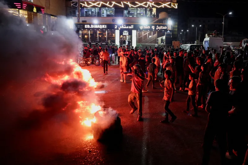 Syria Economic Crisis: Austerity Backfires on Sharaa A nighttime protest near Aleppo, Syria, where a crowd surrounds a large fire burning in the street, demonstrating against rising fuel prices amid the Syria Economic Crisis — a sign of how austerity has backfired on Sharaa.