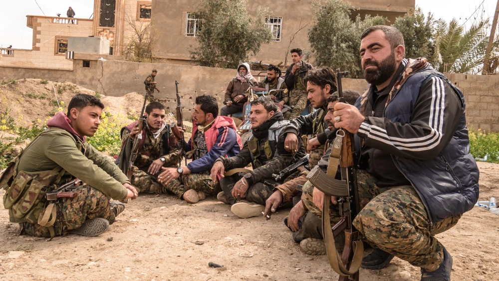 Kurdish Reversal Accelerates as US Abandons Former Allies A dusty outdoor photograph showing a group of Syrian Democratic Forces fighters from Raqqa resting on the ground in Baghouz, holding AK-47 rifles while wearing a mix of camouflage and civilian jackets, symbolizing the demoralizing geopolitical setbacks that define the accelerating Kurdish Reversal as the US abandons its former allies.