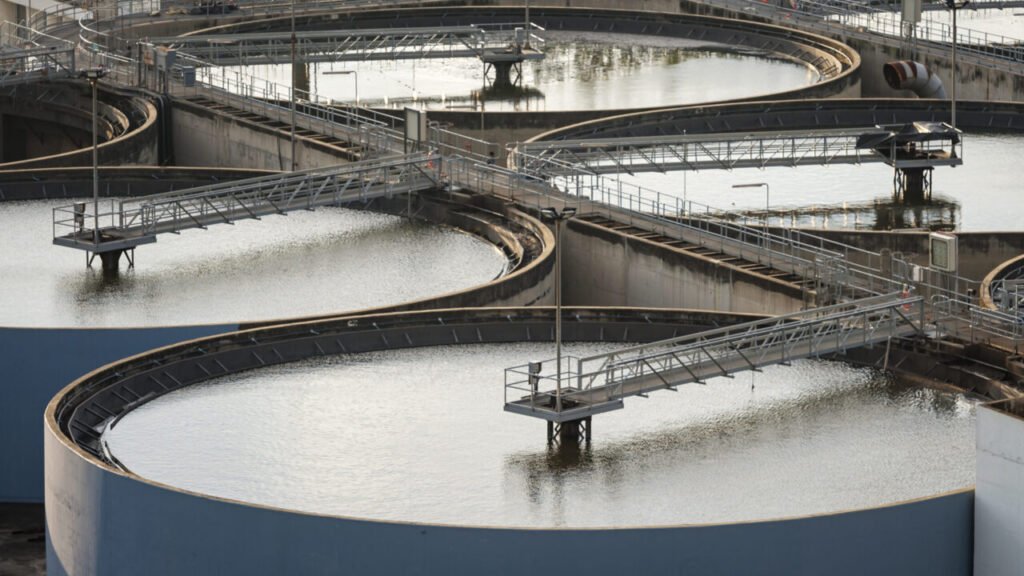 An elevated photograph showing several large circular water treatment clarifier tanks with metal walkways and bridges crossing over the water, visually representing the critical civilian infrastructure that is directly targeted and threatened by the escalating Iran Cyber Strategy against US water utilities.