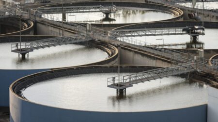 An elevated photograph showing several large circular water treatment clarifier tanks with metal walkways and bridges crossing over the water, visually representing the critical civilian infrastructure that is directly targeted and threatened by the escalating Iran Cyber Strategy against US water utilities.