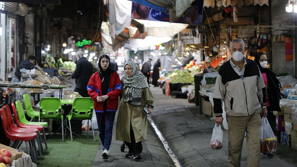 A bustling street market in an Iranian bazaar showing ordinary citizens shopping for groceries, with a man wearing a white mask carrying plastic bags and two women in headscarves walking past produce stalls, visually representing the disproportionate impact of Financial Sanctions on the daily lives of Iran's poor compared to the wealthy elite.