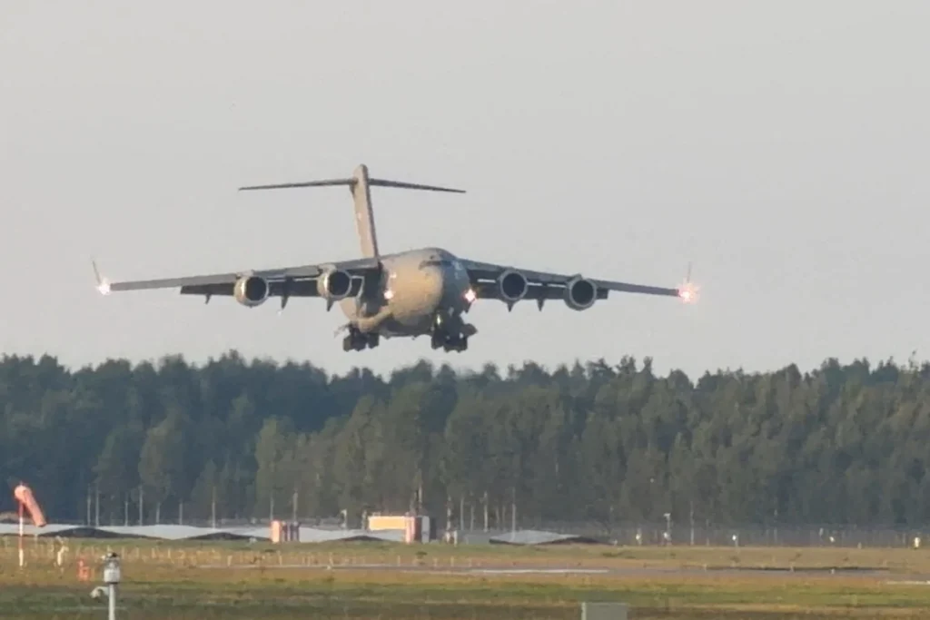 A low-angle photograph showing a US Air Force C-17 Globemaster transport aircraft with its landing gear down and lights illuminated, coming in for a landing at an airfield near Riga, Latvia, carrying CIA Director John Ratcliffe, symbolizing the high-stakes geopolitical stakes of the Ratcliffe Moscow trip that draws a definitive red line on Iran intelligence cooperation.