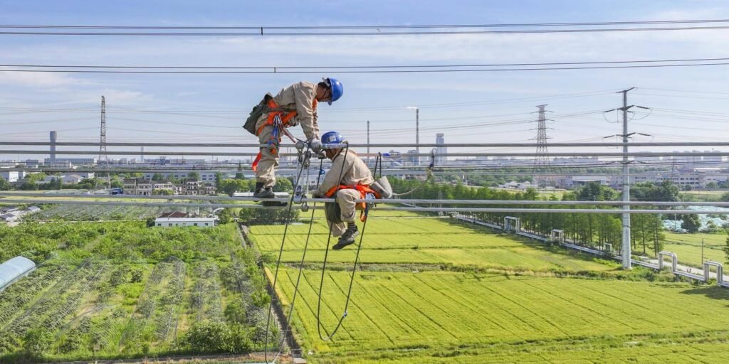 أمن الطاقة في الصين يتجاوز هرمز لكنه يخلق مخاطر Two linemen in orange harnesses and blue hard hats work atop high-voltage transmission lines stretched above lush green fields, with power towers and an industrial skyline fading into the distance — the grid at the heart of China energy security as it weathers Hormuz but creates new risk.