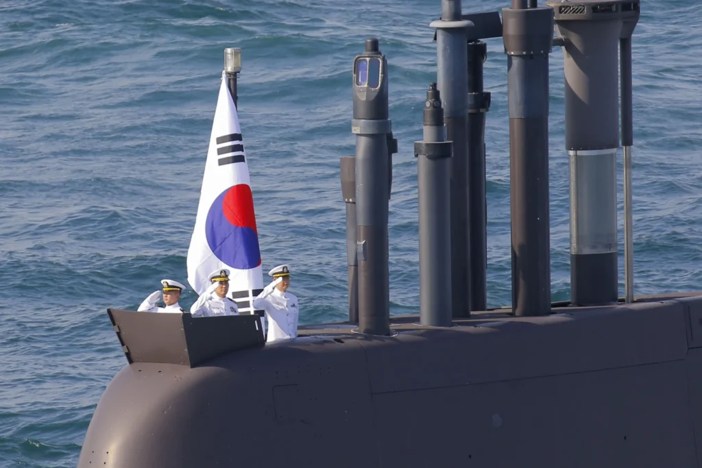 US Allies in Asia: Time to Stop Free-Riding on America Three South Korean sailors in white dress uniforms salute from the conning tower of a submarine flying the South Korean flag, with several periscopes and masts raised above the dark hull, a scene that reflects the naval power of US Allies in Asia.