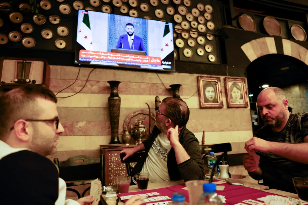 رفع العقوبات عن سوريا يفتح الباب أمام الاقتصاد العالمي A cozy coffee shop scene in Damascus showing men playing cards and drinking tea while a televised address by Syrian President Ahmed al-Sharaa plays in the background, symbolizing the everyday civilian life poised to benefit as Syria sanctions relief opens the door to the global economy.