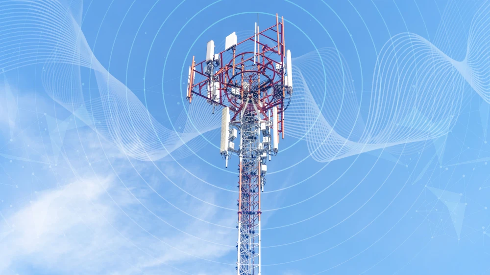 الاتصالات العراقية: قبضة الميليشيات التي تتجاهلها واشنطن A red-and-white lattice telecom tower bristling with antennas rises against a blue sky, overlaid with digital signal waves and a network map — infrastructure at the center of Iraqi Telecom, where a militia grip goes overlooked by Washington.