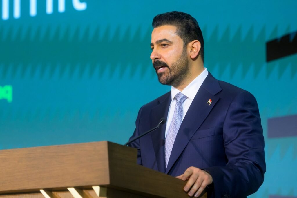 A profile photograph of Iraqi Prime Minister Ali al-Zaidi speaking at a wooden podium at the U.S. Chamber of Commerce in Washington, wearing a dark blue suit and a light blue patterned tie, symbolizing his government's active response to the Iraq Fiscal Shock and his drive for sweeping anti-corruption reforms.