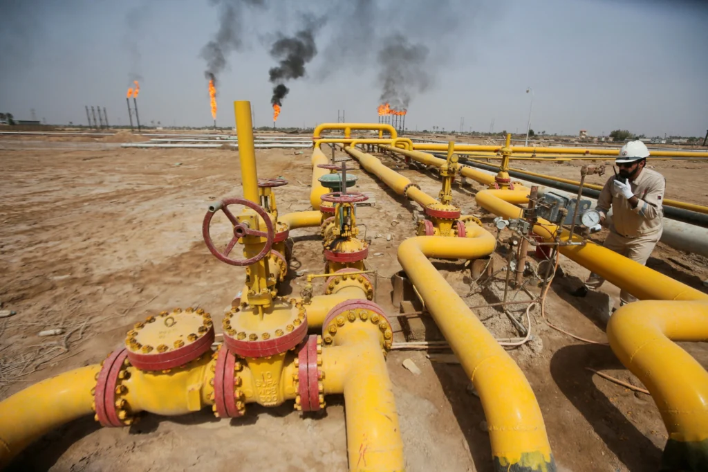 US-Iraq Oil Deals Fail Without Disarming Militias A worker in a white hardhat and uniform inspects yellow pipelines and pressure gauges at a desert oil field with gas flares in the background, highlighting the infrastructure at the heart of US-Iraq Oil Deals that fail without disarming militias.
