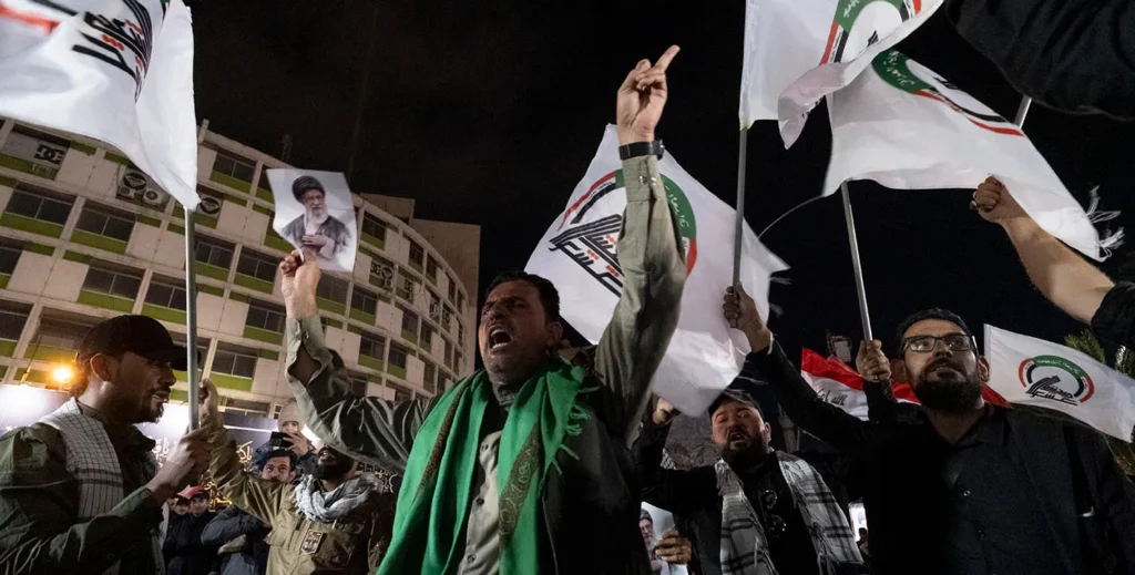 Iraqi PMF reform matters more than the September 30 deadline A nighttime rally with men waving white PMF flags and holding a portrait of Ayatollah Ali Khamenei, one protester shouting with a green scarf over his shoulders, capturing the popular base that any Iraqi PMF reform must navigate.