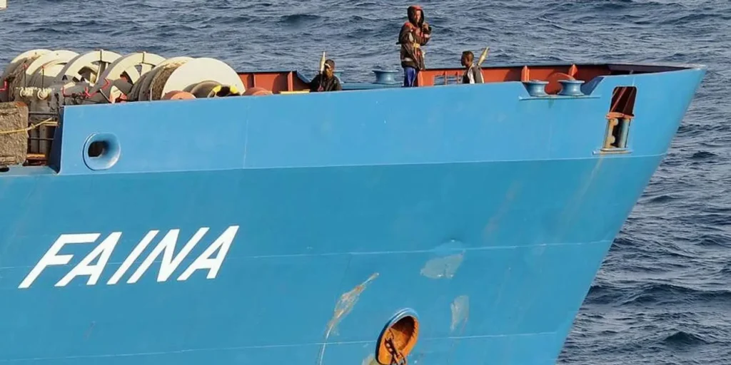 The blue cargo ship FAINA, historically hijacked by Somali pirates in 2008, sails open waters with a small crew on deck, embodying the Somali Piracy Resurgence amid an Iran War vacuum.