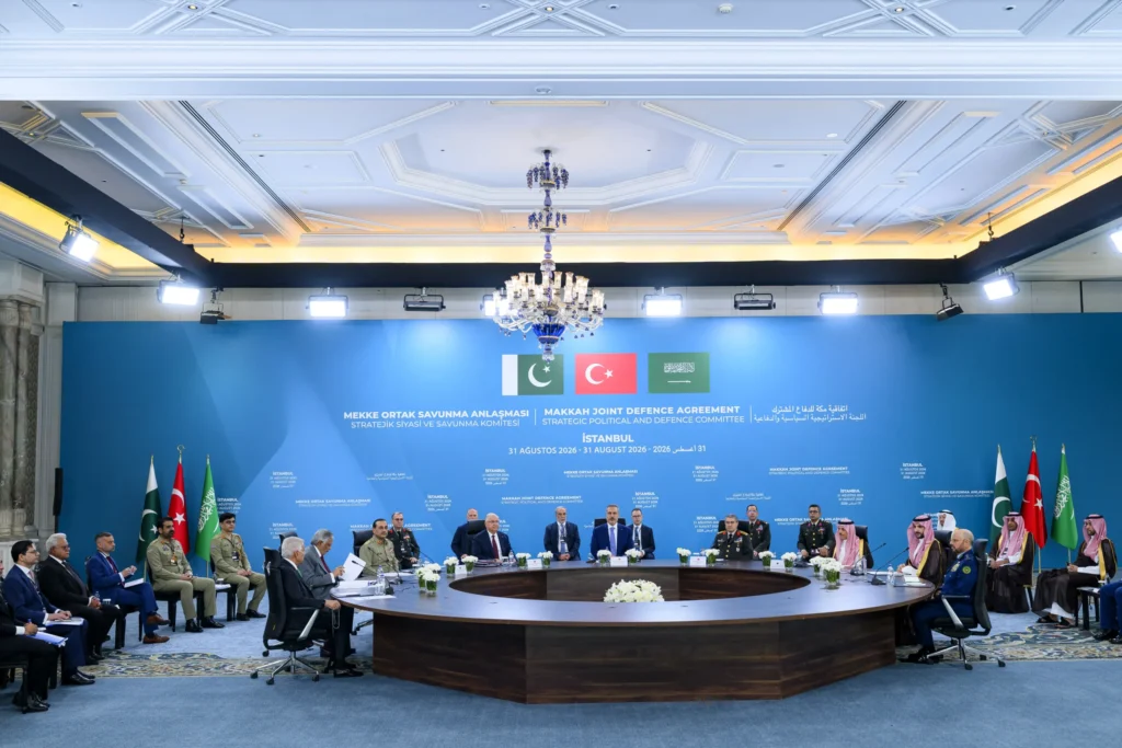 Mecca Alliance Could Anchor a New Regional Order A formal photograph of a high-level summit in Istanbul showing delegates from Turkey, Pakistan, and Saudi Arabia, along with military officers, gathered around a U-shaped table in front of a blue backdrop announcing the "Makkah Joint Defence Agreement," visually representing the strategic political and defense coordination of the Mecca Alliance that could anchor a new regional order.