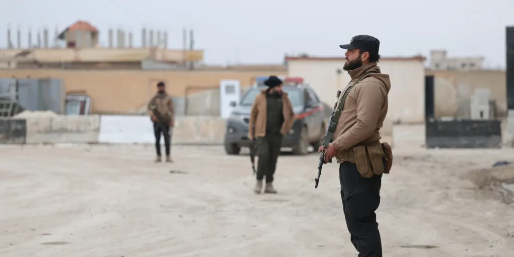 A dusty outdoor photograph showing a bearded man in a tan jacket, black cap, and tactical vest, holding an assault rifle and looking off-camera, with two other armed men and a pickup truck in the blurred background, symbolizing the on-the-ground security presence that underscores why Syrian Kurds require robust decentralization rather than mere integration into central state structures.