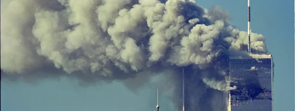 A photograph showing the Twin Towers engulfed in a massive plume of thick grey smoke on September 11, 2001, with the iconic antenna on the North Tower visible against a clear blue sky, symbolizing the catastrophic scale of Mass-Effect Terrorism that now risks returning with new drone and AI technologies.