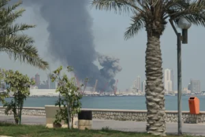 A striking waterfront photograph showing thick, dark smoke billowing from the port and city skyline across the water, framed by a relaxed foreground of palm trees and a stone promenade, vividly illustrating the catastrophic destruction of the Iran War Disaster that severely devalues US military prowess worldwide.