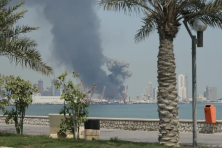 A striking waterfront photograph showing thick, dark smoke billowing from the port and city skyline across the water, framed by a relaxed foreground of palm trees and a stone promenade, vividly illustrating the catastrophic destruction of the Iran War Disaster that severely devalues US military prowess worldwide.