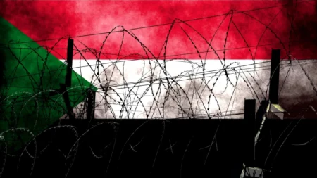 Barbed wire and dark fence posts frame a textured Sudanese flag in the background, capturing a Sudan partition that is emerging without a formal declaration.