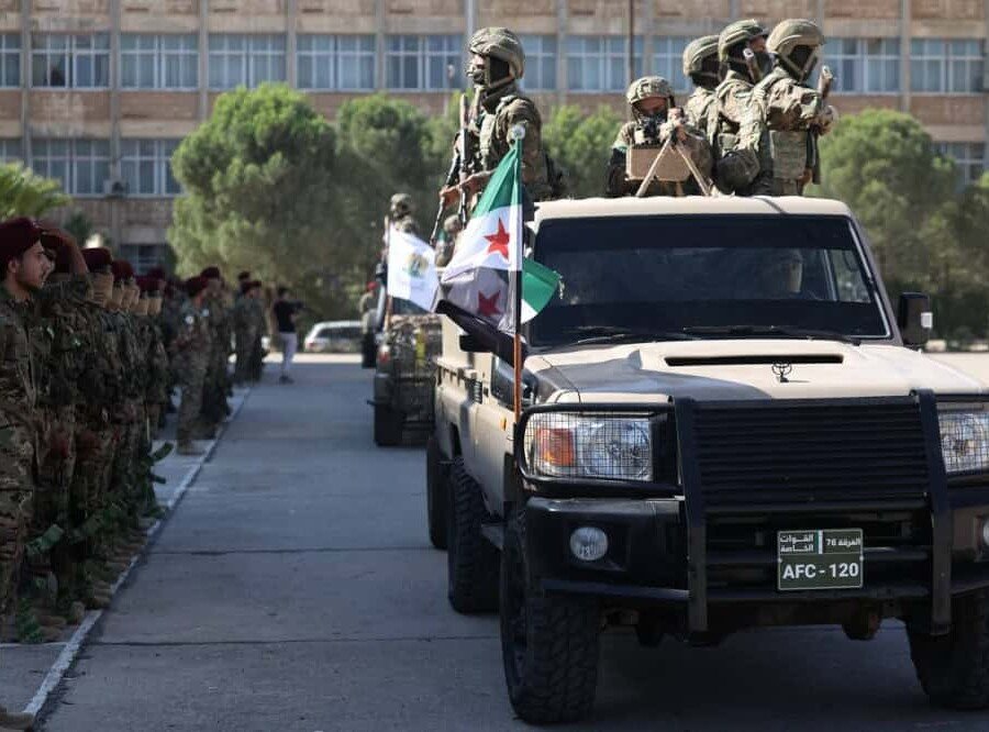 Syrian military absorbs extremists the US designated Heavily armed fighters in tactical gear and face coverings ride atop a military vehicle flying the Syrian independence flag past a formation of soldiers, a scene of the Syrian military absorbing extremists the US designated.