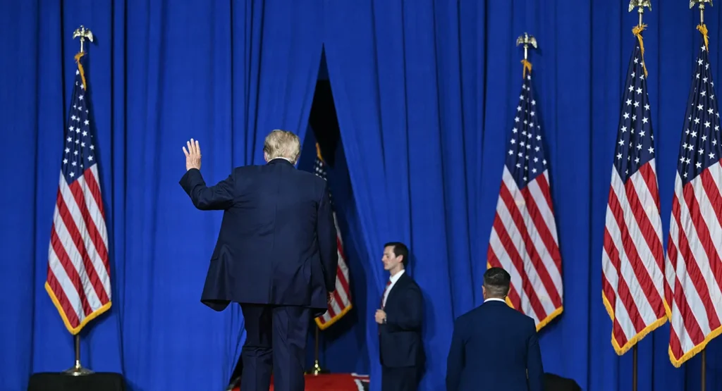 A dramatic photograph showing President Donald Trump from behind as he walks through a parting in blue stage curtains, raising his hand in a wave, flanked by rows of American flags on golden poles, symbolizing the departure from traditional democratic leadership that defines the Trump Democracy retreat, which leaves the world without a champion.