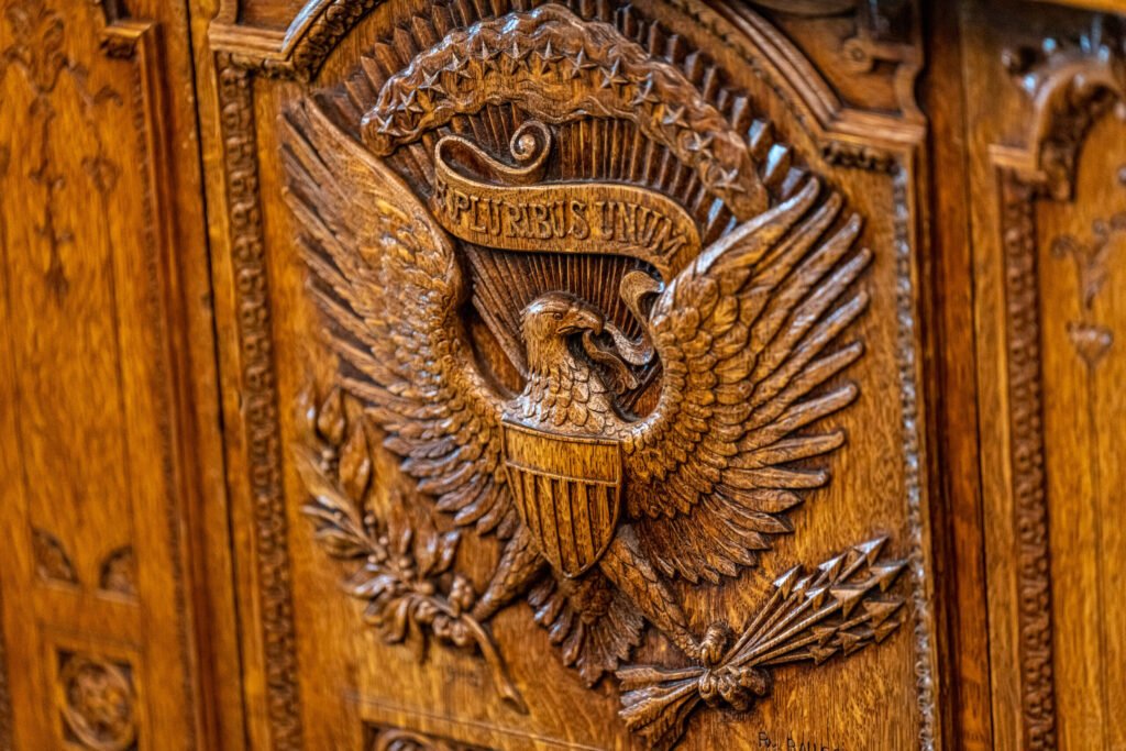 A detailed close-up photograph of the intricately carved wooden Great Seal of the United States on the Resolute Desk, featuring the American bald eagle holding an olive branch and arrows with the "E PLURIBUS UNUM" banner, symbolizing the executive power and diplomatic gravitas required for a strategic Iran Policy that demands smarter planning rather than more force.