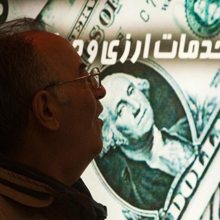 چرا تحریمهای ایران توسط ایالات متحده به بانکداری امارات متحده عربی هدف قرار میگیرد A close-up profile photograph of a man wearing glasses, intently looking up at a glowing digital currency exchange board displaying a large $100 US dollar bill and Persian text, visually illustrating the vital financial channels and daily economic anxiety that US Iran Sanctions aim to disrupt by specifically targeting UAE banking hubs used for dollar transfers and sanctions evasion.