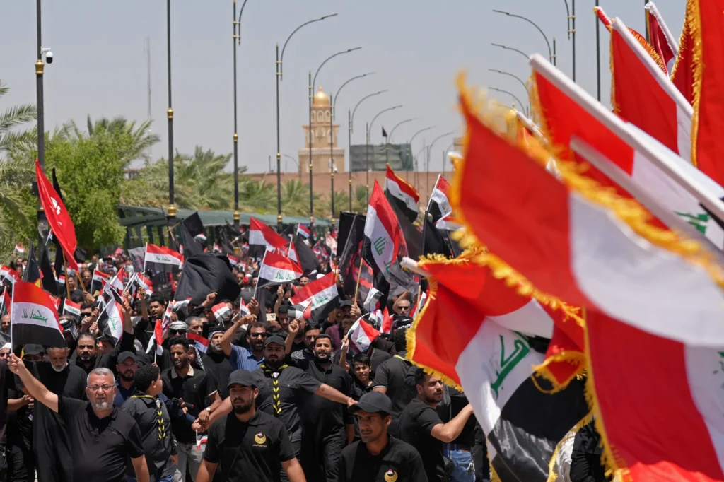 A massive street demonstration in Baghdad showing crowds of men dressed in black marching while waving Iraqi national flags alongside militia banners and black mourning flags, with a golden-domed mosque visible in the background, visually representing the powerful Iran-backed factions whose show of force deepens the Iraq Political Crisis and directly tests Prime Minister Ali al-Zaidi's balancing act.