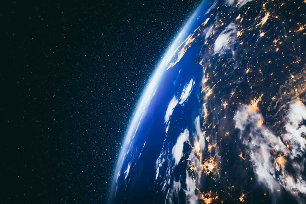 A high-orbit satellite photograph of Earth at night, illuminated by golden city lights beneath scattered clouds and the vibrant blue atmosphere along the horizon, set against the vast blackness of space, symbolizing the interconnected and fragile world order that is currently straining under a deepening Global Crisis.