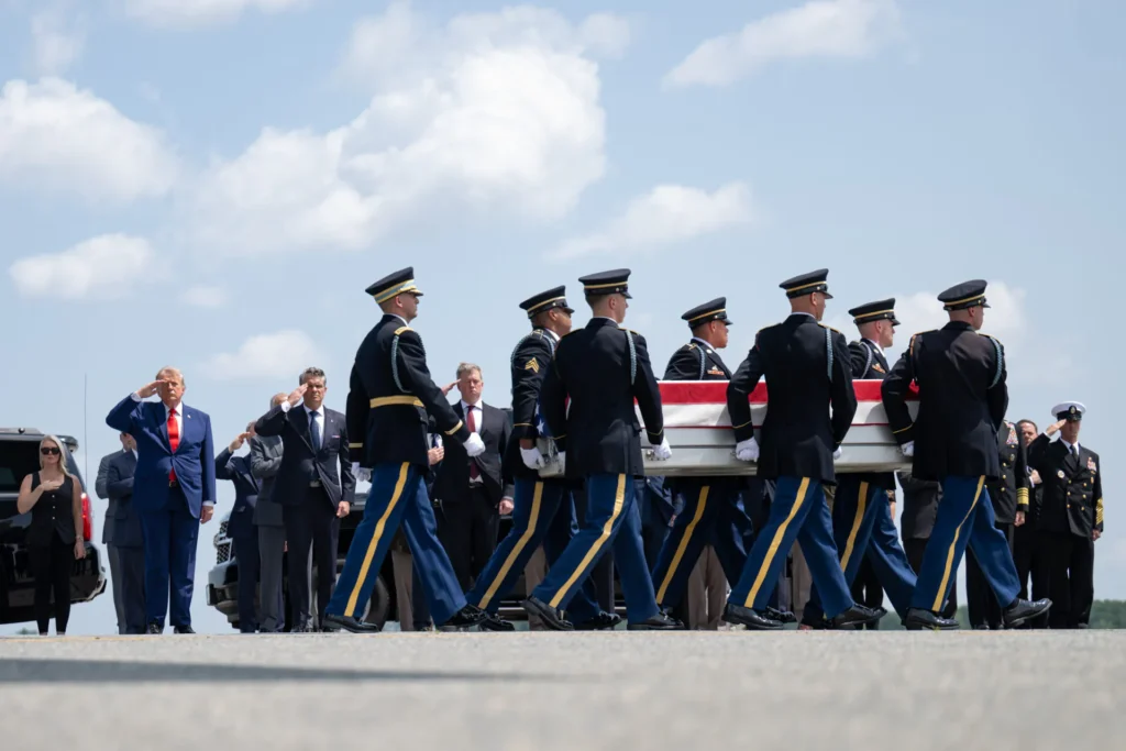 US Overseas Bases Slip From Tripwire to Iranian Bullseye Donald Trump in a blue suit and red tie stands at attention and salutes alongside military officers as an Army honor guard carries a flag-draped casket across an airfield tarmac, a somber scene of the human costs tied to US Overseas Bases.