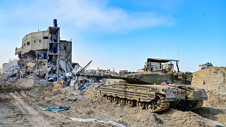 Israeli public opinion gives Netanyahu political cover A photograph of an Israeli Merkava tank driving through the rubble of a heavily destroyed building in Gaza under a clear blue sky, visually representing the ongoing military campaign that continues to be sustained by the Israeli public opinion providing political cover for Prime Minister Netanyahu.