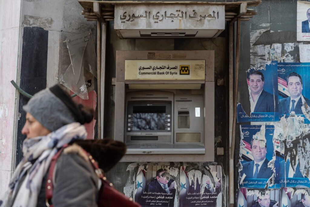 A woman walks past a darkened ATM of the Commercial Bank of Syria, flanked by torn political posters on a battered street, highlighting the struggle for Syria reconstruction and the critical need to revive state banks.