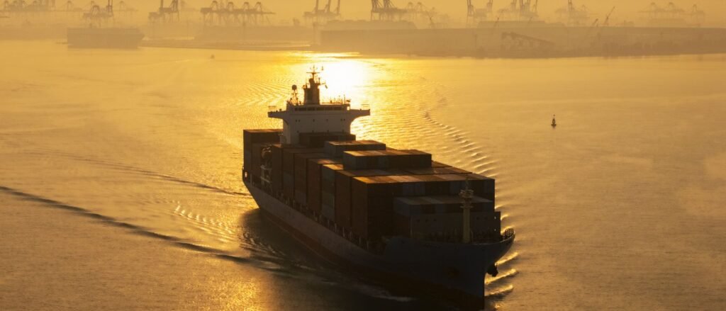 Hormuz Corridor Deal Locks In Iran’s Chokehold A golden-hour photograph of a large container ship navigating through hazy waters with industrial port cranes silhouetted in the background, symbolizing the vital maritime trade route at the center of the Hormuz Corridor Deal that effectively locks in Iran's strategic chokehold over global shipping lanes.