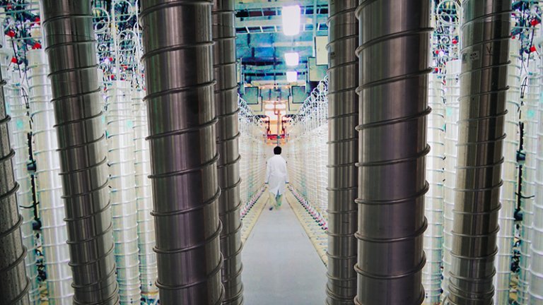 قدرت چانهزنی هستهای ایران با وجود شکست نظامی حفظ میشود A deep-perspective photograph showing a central aisle flanked by rows of tall, silver gas centrifuge cascades and complex piping in a sterile enrichment facility, with a scientist in a white coat walking away down the corridor, symbolizing the enduring industrial capacity that preserves Iran Nuclear Leverage in the face of significant military defeat.