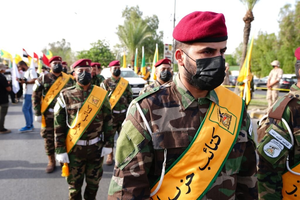 لماذا تواجه نزع سلاح الميليشيات العراقية مقاومة شديدة A photograph showing uniformed Iraqi militia fighters wearing red berets, black face masks, and yellow ceremonial sashes marching in a disciplined formation, visually demonstrating the organized armed defiance that fuels the heavy resistance against Iraq Militia Disarmament initiatives.