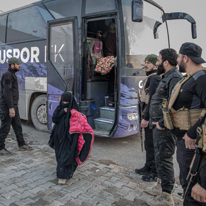 محاكمات داعش تتجاوز المهمة العسكرية الأمريكية في العراق A woman in a black niqab steps off a purple bus carrying a bright pink jacket, surrounded by armed men with US flag patches in Iraq, where ISIS Trials outlast the US military mission.