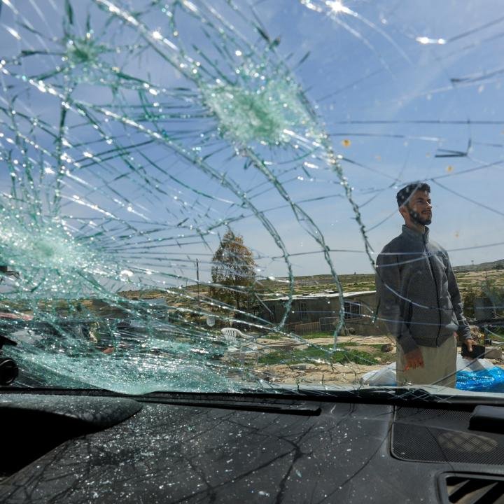 Settler violence: a systemic failure, not a policing gap Seen through a shattered windshield with multiple impact craters, an Israeli settler in a grey jacket stands in an arid landscape, a stark aftermath of Settler violence that reflects a systemic failure, not a policing gap.