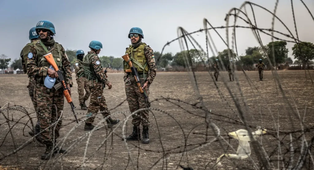 UN peacekeepers in blue helmets and camouflage uniforms stand with rifles in a dry open field behind coils of barbed wire in South Sudan, a scene that embodies the UN Decomposition driven by US selectivity.