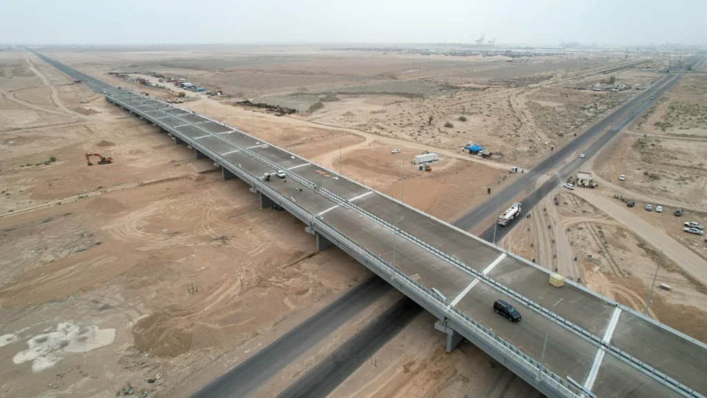 طريق التنمية العراقي يواجه خطر التحول إلى ساحة عقود An aerial view of a newly built concrete bridge and highway cutting across an arid desert landscape, with construction vehicles, workers in safety vests, a black SUV driving along the overpass, and a distant port with cranes visible on the hazy horizon — a scene of large-scale infrastructure taking shape along the Iraqi Development Road, which risks becoming a contract arena.