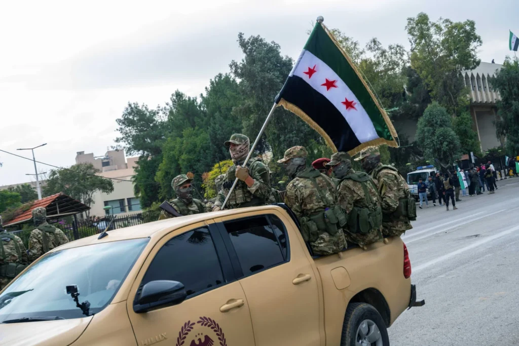 Syria suspicious deaths revive Assad-era purge fears Armed men in camouflage uniforms and face coverings ride in a tan pickup truck flying the Syrian independence flag, highlighting the tense atmosphere where Syria suspicious deaths revive Assad-era purge fears.