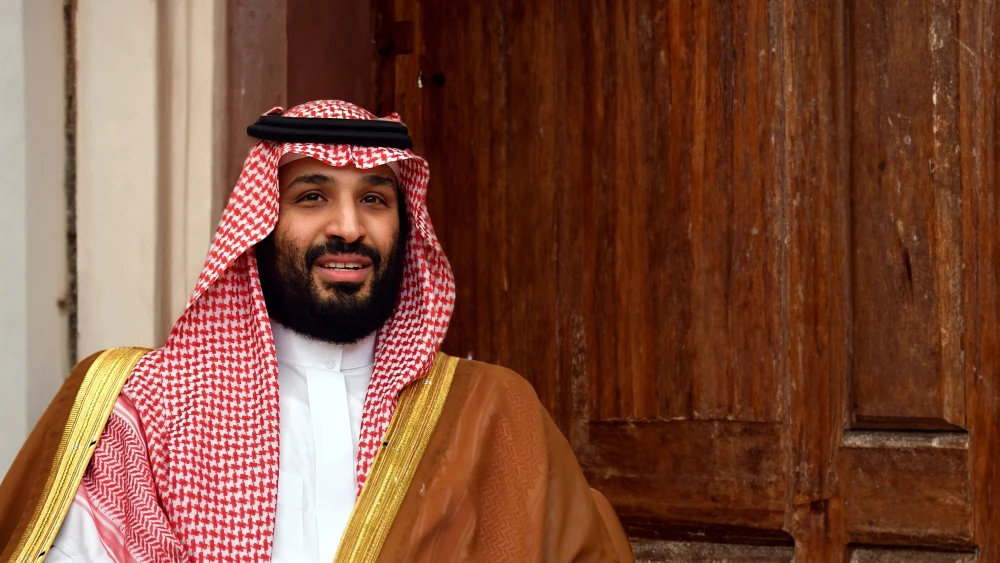 MBS Rivalry With UAE Threatens Riyadh’s Control Over Yemen A portrait of Saudi Crown Prince Mohammed bin Salman wearing a red-and-white shemagh and a brown bisht, smiling slightly in front of a wooden door. His composed expression contrasts with the tense regional dynamics where the MBS Rivalry with the UAE threatens Riyadh's control over Yemen.