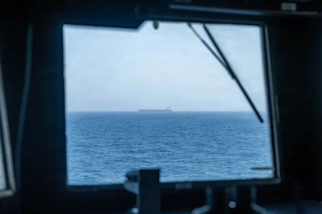 Viewed through the dark window of a ship's bridge, a lone tanker crosses the open sea on the horizon — a quiet image of the global oil trade that a Hormuz closure would disrupt, proving US oil independence is a myth.