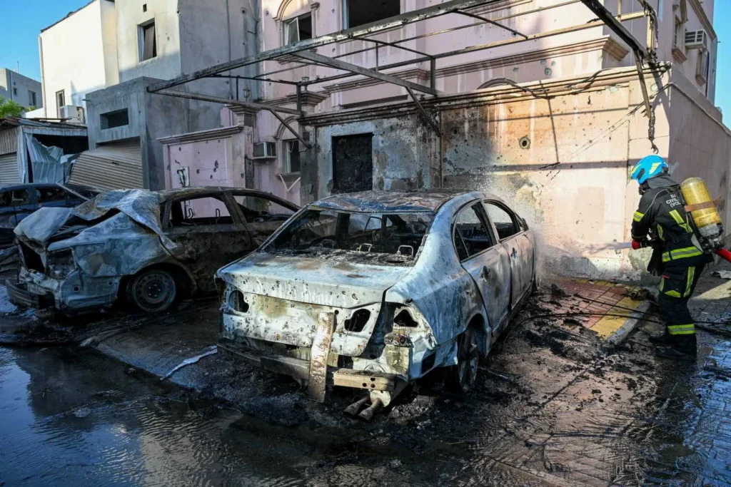 Gulf Economic Quagmire Emerges as Iran War Drags On A street-level photograph showing two charred, completely burned-out cars parked beside a heavily damaged residential building with visible shrapnel marks, as a firefighter in protective gear and breathing apparatus surveys the wreckage on the wet pavement, visually representing the physical destruction and escalating chaos that deepens the Gulf Economic Quagmire as the Iran war drags on.