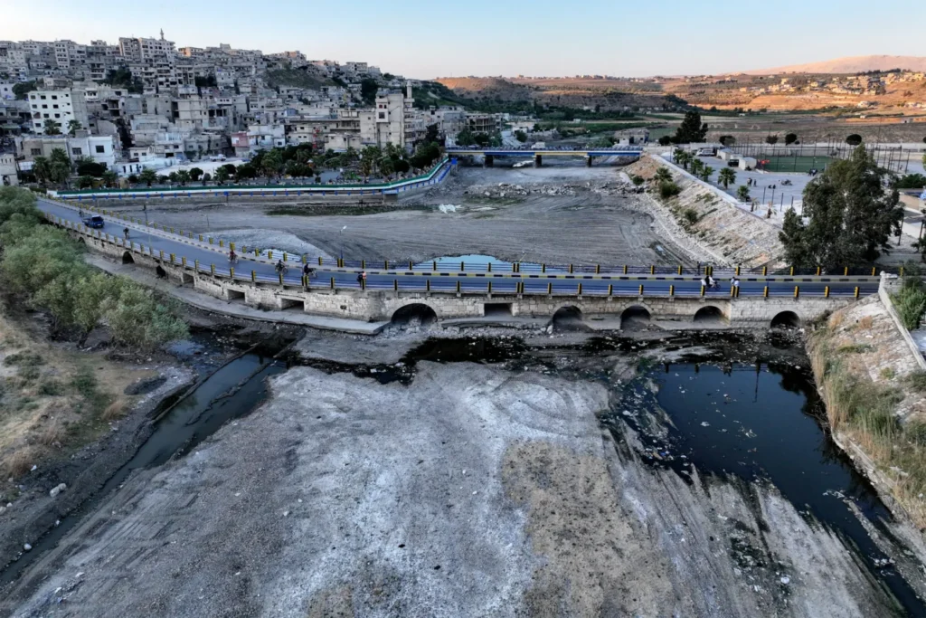 بازسازی سوریه، جبهه فراموششده جنگ ایران A nearly dry riverbed littered with debris runs beneath a stone arch bridge and a curving riverside road, with dense concrete housing climbing the hillside behind — a landscape that captures the stalled Syria recovery on the Iran war's forgotten front.