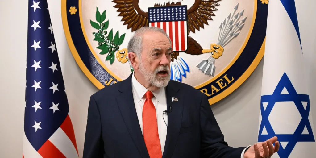 West Bank settler violence tests US accountability tools A close-up photograph of US Ambassador to Israel Mike Huckabee speaking at a diplomatic event, set against the official US Embassy seal and flanked by the US and Israeli flags, visually representing the diplomatic pressure points where West Bank settler violence tests the limits of US accountability tools.