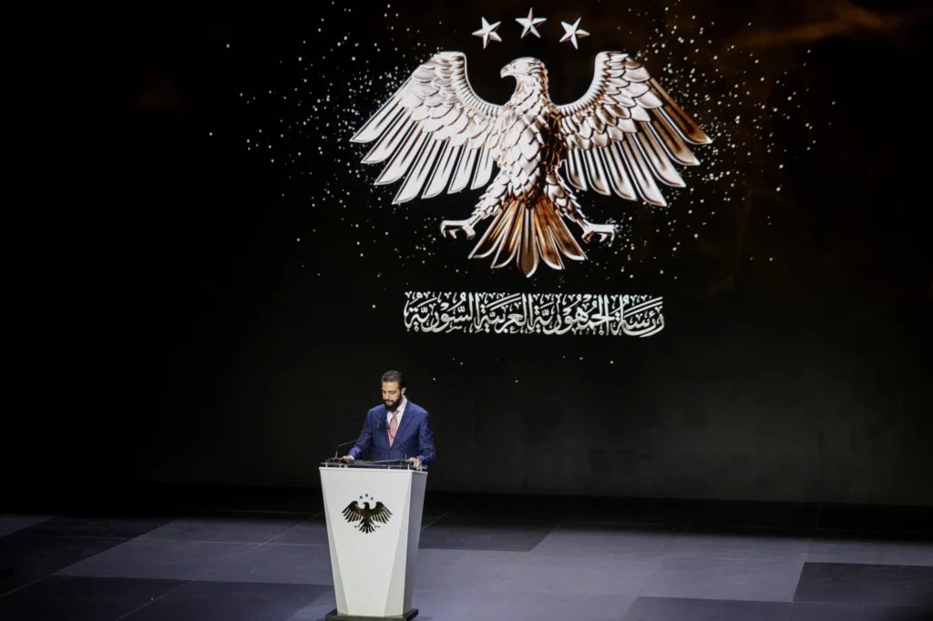 Syria Policy: Won Against ISIS, Lost the Country Ahmed al-Sharaa, wearing a blue suit and red tie, speaks at a podium bearing the Syrian eagle emblem, with a large screen behind him displaying the same eagle and three stars above Arabic text — a formal address that captures the paradox of Syria Policy: winning against ISIS while losing the country.