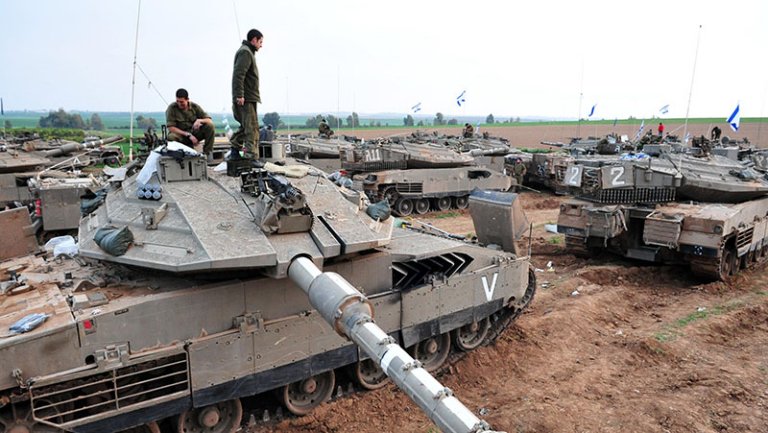 Israeli Occupation Reflects Policy, Not Wartime Necessity Israeli Merkava tanks parked in a muddy field with soldiers and Israeli flags, illustrating the Israeli Occupation that reflects policy, not wartime necessity.