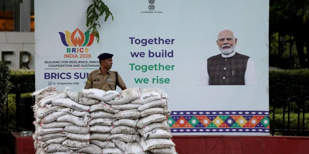 A security officer stands behind a stack of cement bags in front of a large promotional billboard for the BRICS Summit 2026 in India, featuring Prime Minister Narendra Modi and the slogan "Together we build, Together we rise," symbolizing the upcoming diplomatic gathering that is being tested by the Iran war splitting the bloc and India's challenge to hold it together.