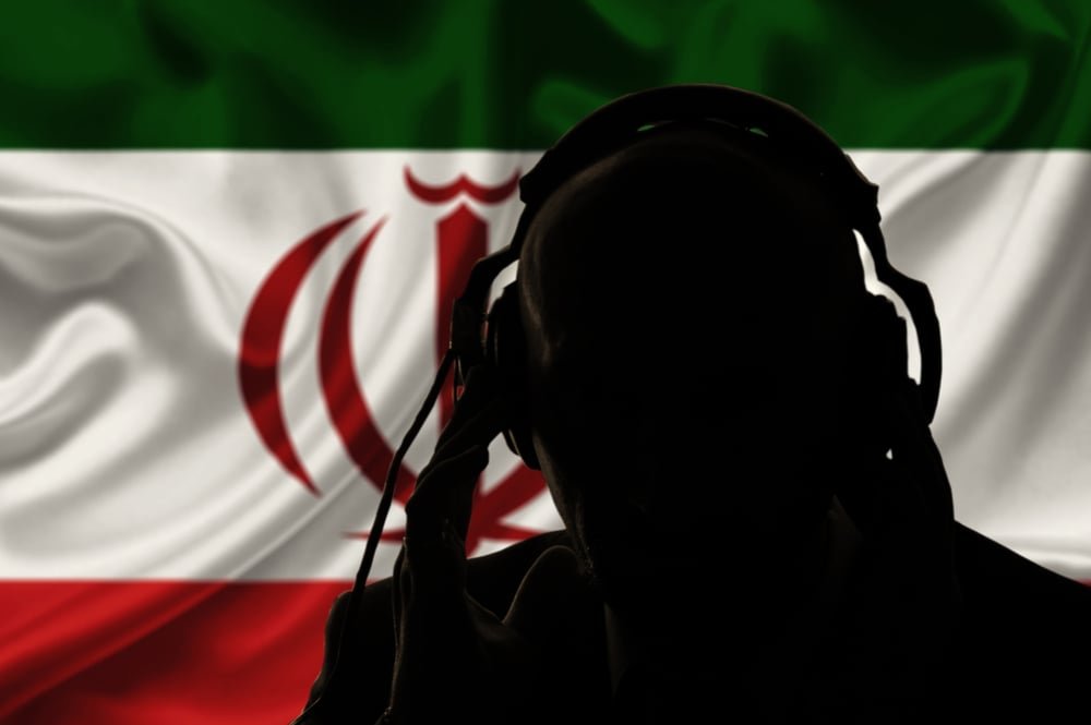 Iran Transnational Repression Turns Data into Hit Lists A silhouetted figure wearing large headphones stands before a blurred Iranian flag, evoking the surveillance and monitoring that define Iran Transnational Repression.