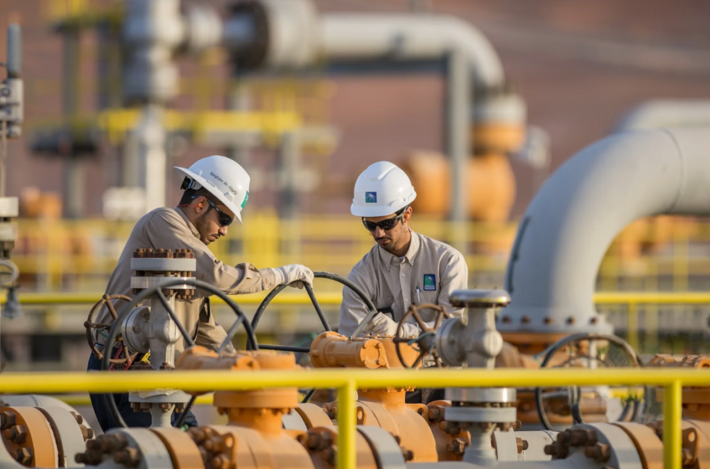 Two Saudi Aramco employees in white hard hats turn a valve wheel among orange and yellow pipelines at an oil facility, as Gulf bypass routes turn redundancy into rationing.