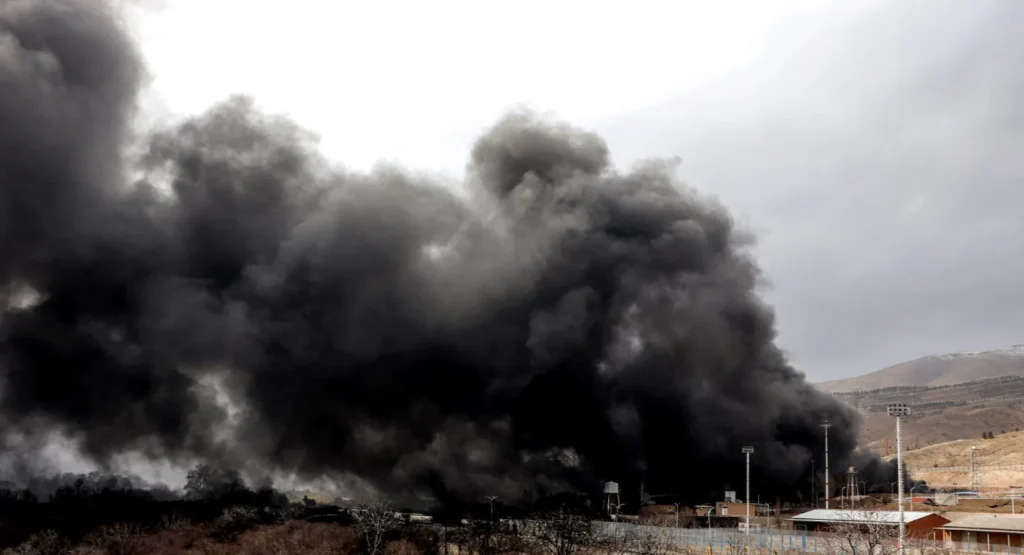 پیامدهای جنگ ایران: تداخل اقلیم، بهداشت و حکومت Thick black smoke billows over an arid landscape and infrastructure, symbolizing the Iran War Fallout where climate, health, and governance collide.