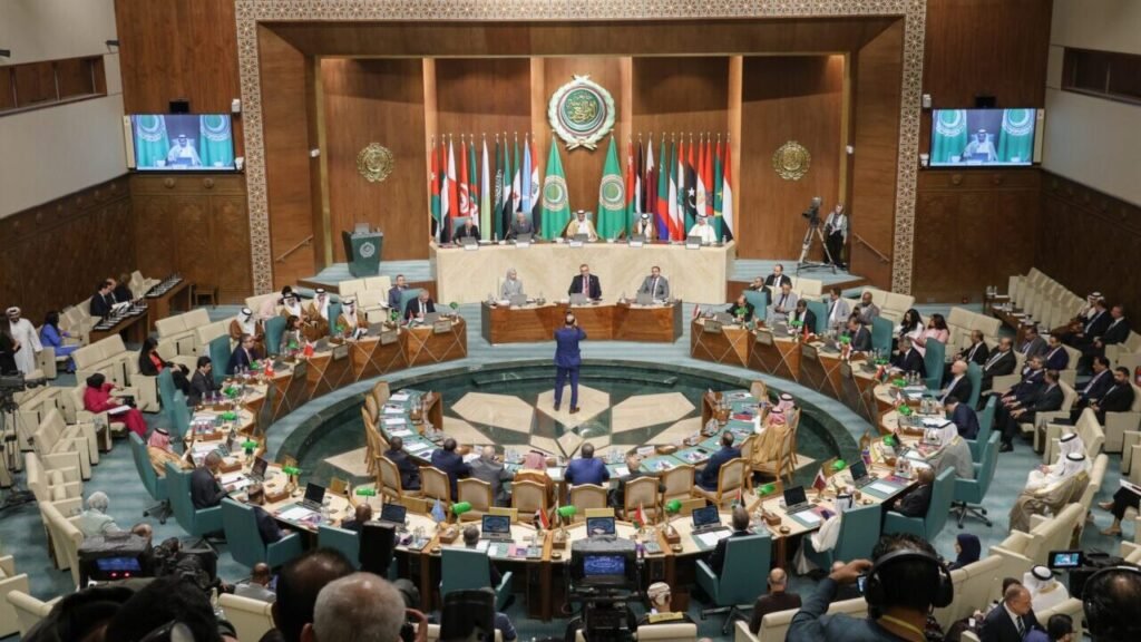 A wide-angle photograph of an Arab League summit showing delegates seated in a large circular formation around a central emblem, with rows of member state flags displayed behind the main podium, visually representing the collective multilateral engagement of Arab Diplomacy that offers the US a smarter strategic path in the Middle East.